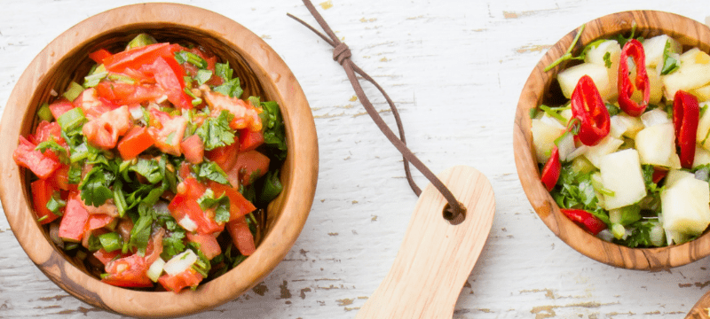 Three wooden bowls with arugula, diced tomatoes, and pineapple mix. Cutting board with cucumbers and knife on the right.
